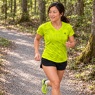 YUL Neon Radioactive Pine women's running shirt, neon yellow, worn by a runner on a wooded path.